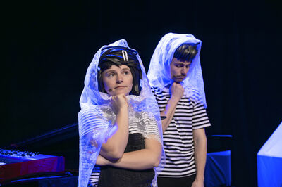 Twee performers staan naast elkaar op een donker podium, beiden met een witte, doorschijnende sluier over hun hoofd. Ze dragen gestreepte shirts; de persoon vooraan heeft een tuinbroek aan en kijkt peinzend voor zich uit met een hand onder de kin, terwijl de ander iets achter haar staat in een vergelijkbare nadenkende houding. Het blauwe podiumlicht en een keyboard op de achtergrond versterken de theatrale sfeer.