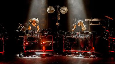 Twee percussionisten spelen op uitgebreide drumopstellingen op een donker podium. Hun lange haren zwaaien mee terwijl ze energiek slaan op trommels en bekkens. Spotlights van boven verlichten hen, met rood licht onder de instrumenten, wat zorgt voor een krachtige en dynamische concert­sfeer.