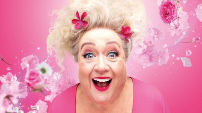 Christel de Laat, haar versierd met roze bloemen, lacht enthousiast met open mond tegen een felroze achtergrond vol zwevende bloesems en rozen.