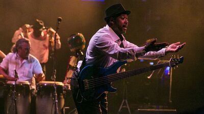 Een bassist met een donkere hoed en wit overhemd staat op een podium en maakt een expressief gebaar met zijn handen, terwijl zijn basgitaar om zijn schouder hangt. Achter hem speelt een percussionist op conga’s en zijn andere bandleden vaag zichtbaar in warm podiumlicht. De sfeer is energiek en muzikaal.