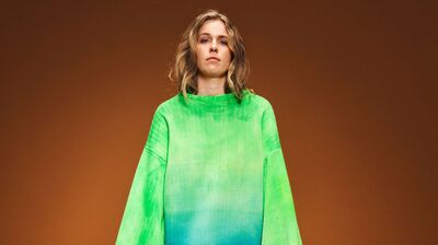 Model in een oversized groene trui met verloop van helder naar diepgroen, tegen een warme oranje achtergrond.