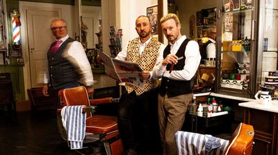 Drie mannen in een kapsalon staan naast elkaar, gekleed in klassieke outfits, met een krant in de hand.