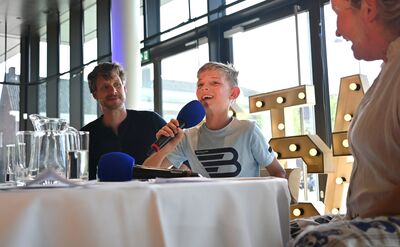 Jongentje met een microfoon praat tijdens een interview voor een publiek, met volwassenen op de achtergrond.