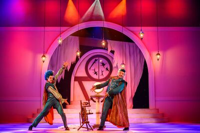 Twee acteurs in opvallende, kleurrijke kostuums staan op een fel verlicht podium met roze en paarse tinten. Ze dragen lange mantels en maken theatrale gebaren naar een gloeiende bol op een sokkel tussen hen in. Op de achtergrond is een grote boog met een ronde deur en een klok te zien, terwijl meerdere hangende lantaarns boven het toneel hangen.