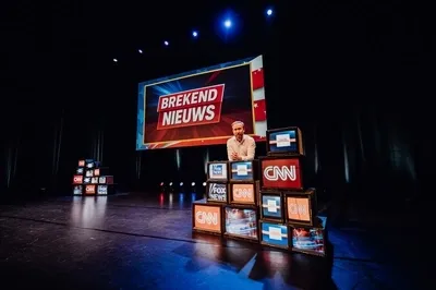  Raymond Mens in een wit overhemd staat op een donker podium achter een opstapeling van oude televisies. Op de televisieschermen zijn logo's te zien van CNN en FOX News. Op een groot projectiescherm achter hem staat de tekst 'BREKEND NIEUWS' tegen een rood-blauwe achtergrond met witte sterren.