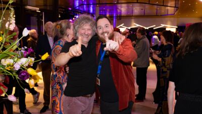 Twee mannen poseren lachend en geven met hun vingers het nummer één aan op een feestelijke bijeenkomst.