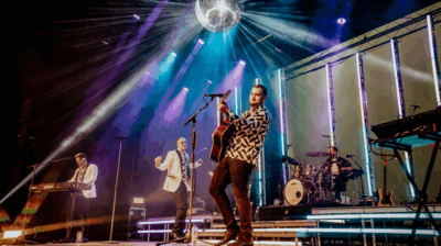 Een band treedt op op een verlicht podium met kleurrijke spotlights en een spiegelbol aan het plafond. Een gitarist staat vooraan bij een microfoon, terwijl een zanger danst in het midden. Links speelt een toetsenist en achteraan zit een drummer. De sfeer is energiek en feestelijk, met lichtstralen die door de zaal schijnen.