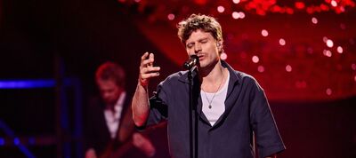 Zanger optredend op een podium met een microfoon, verlicht door kleurrijke lichten op de achtergrond.