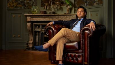 Een man in een donkerblauw pak zit ontspannen in een leren fauteuil in een elegante kamer.