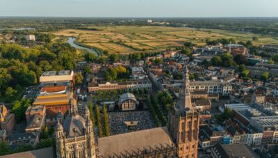 Luchtfoto van een stad met groene velden op de achtergrond en historische gebouwen, inclusief torens en bomen.