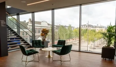 Moderne zithoek met vier donkergroene fluwelen fauteuils rond een gouden tafel, voor een grote glazen pui met uitzicht op De Parade.