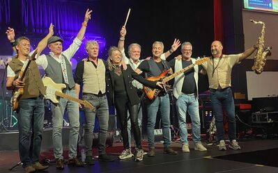 Een band staat op het podium, juichend en poseert met instrumenten na een optreden.