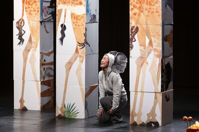 Een performer in een grijs kostuum met grote olifantenoren knielt op een podium tussen hoge, stapelbare zuilen met illustraties van giraffenpoten. De performer kijkt glimlachend omhoog. Het decor heeft een speelse, dierentuinachtige uitstraling met jungle-elementen.