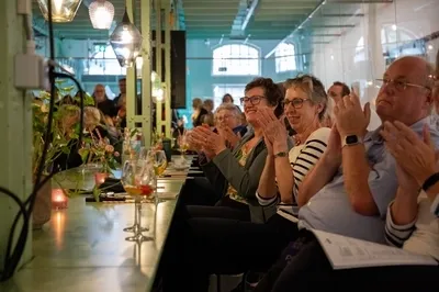 Een zittend publiek aan lange tafels klapt enthousiast in een sfeervol verlichte, industriële ruimte.