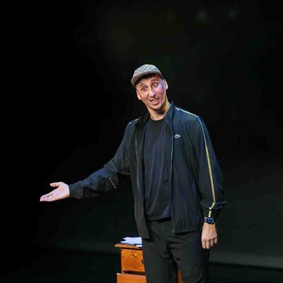 Jawad Es Soufi met pet en zwarte sportieve kleding staat op een donker podium, kijkt met een speelse uitdrukking naar het publiek en maakt een presenterend gebaar met één hand; naast hem staat een klein houten kastje.