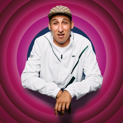 Jawad Es Soufi met pet en witte sportieve jas leunt met zijn armen naar voren tegen een roze achtergrond met concentrische cirkels en kijkt met een licht verbaasde, speelse uitdrukking in de camera.