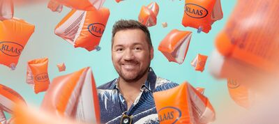 Portret van Klaas van Eerden in een blauw overhemd met een wit golfpatroon. Hij staat centraal in het beeld en wordt omringd door talloze zwevende, oranje zwembandjes waarop de naam 'KLAAS' in een blauw logo staat. De achtergrond is egaal lichtblauw.