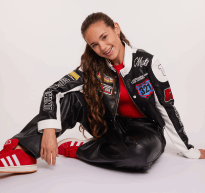 Luna Sabella zit op de grond tegen een lichte achtergrond. Ze draagt een rood shirt onder een zwart-witte jas met verschillende patches en teksten, een zwarte broek en rode sneakers met witte strepen. Haar lange, krullende haar valt over haar schouder terwijl ze ontspannen en vrolijk in de camera kijkt.