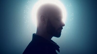 Profiel van een man in silhouet tegen een blauwgroene achtergrond. Rond zijn hoofd straalt een fel wit licht dat een halo-effect creëert, met subtiele lichtpuntjes eromheen. De afbeelding heeft een rustige, bijna spirituele en contemplatieve sfeer.