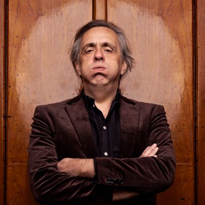 Marcel van Roosmalen met halflang grijs haar staat met over elkaar geslagen armen voor een houten wand, draagt een donker fluwelen jasje en kijkt recht in de camera met een serieuze, licht vermoeide uitdrukking.