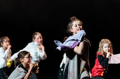 Verschillende kinderen spelen een scène op een donker podium. Een meisje vooraan houdt een kledingstuk over haar schouder en kijkt expressief opzij, terwijl andere kinderen achter haar een vinger voor hun mond houden in een “ssst”-gebaar. Ze dragen verschillende kostuums en hebben geschminkte wangen.