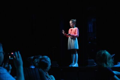 Een jong meisje staat alleen in de spotlights op een donker podium. Ze draagt een roze vest, een lichte rok, kniekousen en witte schoenen. Met haar handen licht opgeheven lijkt ze te spreken of iets uit te beelden. Onderaan in beeld zijn silhouetten van toeschouwers te zien, wat wijst op een optreden of voorstelling.