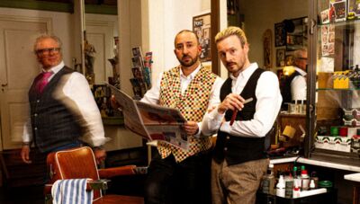 Drie mannen in een kapsalon, gekleed in stijlvolle vesten, poseren met een krant en een scheermes.