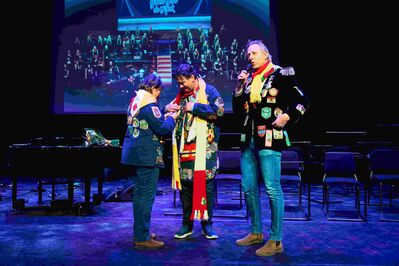 Drie mensen op een podium tijdens een evenement, met kleurrijke jassen en een achtergrond van een podiumgroep.