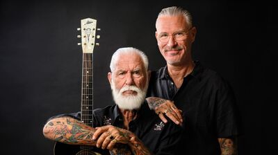 Een portret tegen een zwarte achtergrond van twee mannen die op elkaar lijken. De voorste, oudere man heeft een volle witte baard en zwaar getatoeëerde armen; hij leunt op een akoestische gitaar van het merk Atkin. De jongere man staat achter hem met een hand op zijn schouder, draagt een bril en glimlacht vriendelijk naar de camera. Beiden dragen zwarte overhemden.
