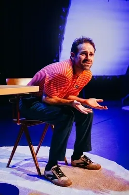 Martijn Crins zit voorovergebogen op een houten stoel op een verlicht rond vloerkleed. Hij draagt een rood-wit gestreept poloshirt en een donkere broek, en kijkt met een vragende, bijna wanhopige blik omhoog terwijl hij zijn handen openhoudt. Op de achtergrond is een groot, blauw verlicht scherm zichtbaar op een verder donker podium.