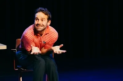 Martijn Crins zit voorovergebogen op een stoel op een donker podium. Hij draagt een oranje-wit gestreept poloshirt en kijkt met een komische, overdreven verbaasde blik opzij terwijl hij zijn handen vragend openhoudt.