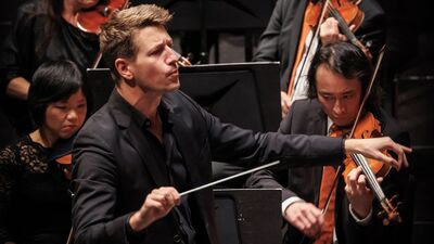 Dirigent met een serieuze blik met violisten achter hem