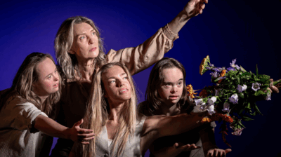 Vier personen poseren dicht bij elkaar tegen een blauwe achtergrond. Eén persoon wijst omhoog terwijl de anderen in dezelfde richting kijken; vooraan houdt iemand een kleurrijk boeket bloemen naar voren. Hun gezichten zijn expressief belicht, met aandacht voor hun blikken en handgebaren.