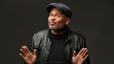 Roue verveer met pet en zwarte leren jas houdt beide handen omhoog met open handpalmen en kijkt opzij met een speelse, afwachtende uitdrukking tegen een donkere achtergrond.