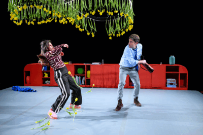 Twee acteurs staan op een theaterpodium met een zwarte achtergrond en een felrode kast achter hen. Boven het podium hangen tientallen gele tulpen ondersteboven aan lange stelen. De vrouw links deinst achteruit terwijl er tulpen op de grond vallen; de man rechts, in een lichtblauw overhemd en stropdas, draait zich naar haar toe met een jas in zijn hand. De scène oogt dynamisch en licht absurdistisch.