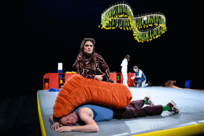 Theatrale scène op een podium met zwarte achtergrond en een rode kast achteraan. Op de voorgrond ligt een man op zijn buik op een mat, deels bedekt door een groot oranje kussen, terwijl een vrouw boven hem zit en ernstig voor zich uit kijkt. Op de achtergrond zijn nog twee acteurs zichtbaar, en boven het podium hangen slingers van gele tulpen. De setting oogt absurdistisch en dramatisch.
