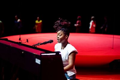 Vrouw speelt piano en zingt op een podium met een rode achtergrond, omringd door andere mensen.
