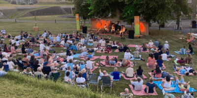 Een buitenconcert met een grote menigte op een grasveld, bezoekers zitten op matjes.
