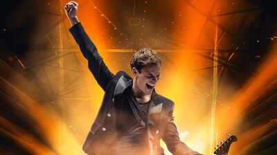 Een man staat op een podium in fel oranje tegenlicht en steekt juichend één arm omhoog. Hij draagt een donker jasje en lijkt midden in een energiek optreden te zitten. Onderaan is een deel van een gitaar zichtbaar, terwijl rook en lichtstralen zorgen voor een krachtige, concertachtige sfeer.