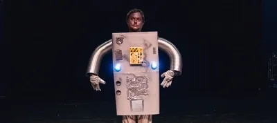 Diederik Jekel staat op een donker podium in een creatief, zelfgemaakt robotkostuum. Het kostuum bestaat uit een grote grijze doos als romp, versierd met knoppen, lampjes en computeronderdelen, en zilveren flexibele buizen als armen. Zijn gezicht is grijs geschminkt en hij kijkt met een serieuze, bijna wezenloze blik vooruit.