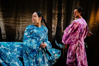 Twee performers staan op een podium met donkere, glanzende gordijnen op de achtergrond. Links draagt een vrouw een wijde, blauw gemarmerde jurk die uitwaaiert terwijl ze glimlachend opzij kijkt. Rechts staat een man in een paars gemarmerd kostuum met volumineuze mouwen, een viool in zijn hand, en kijkt met een lichte glimlach achterom. De belichting geeft de scène een warme, theatrale sfeer.