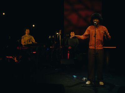 Een zanger staat vooraan op een donker podium en zingt in een microfoon, terwijl hij zijn handen expressief uitstrekt. Hij draagt een warme trui en een grote muts. Links speelt een toetsenist op een keyboard en op de achtergrond zit nog een muzikant. De belichting is zacht en warm, wat zorgt voor een intieme sfeer.